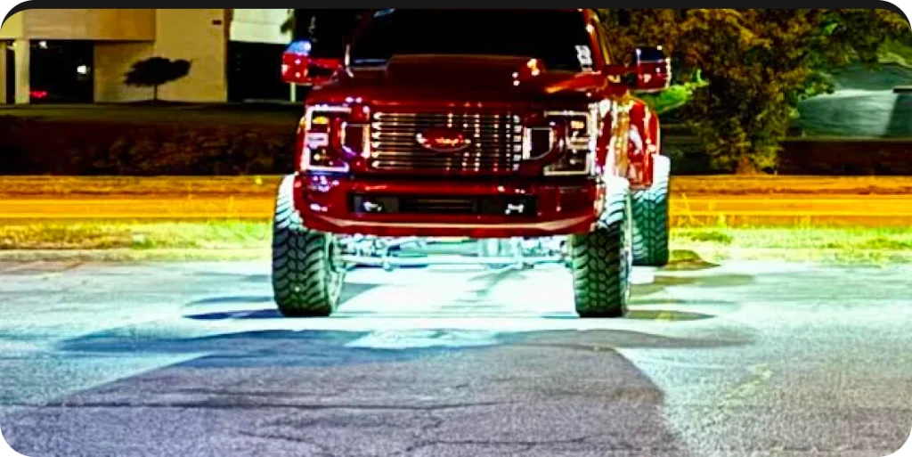 A red, lifted truck with illuminated undercarriage is parked in a lit-up area at night.