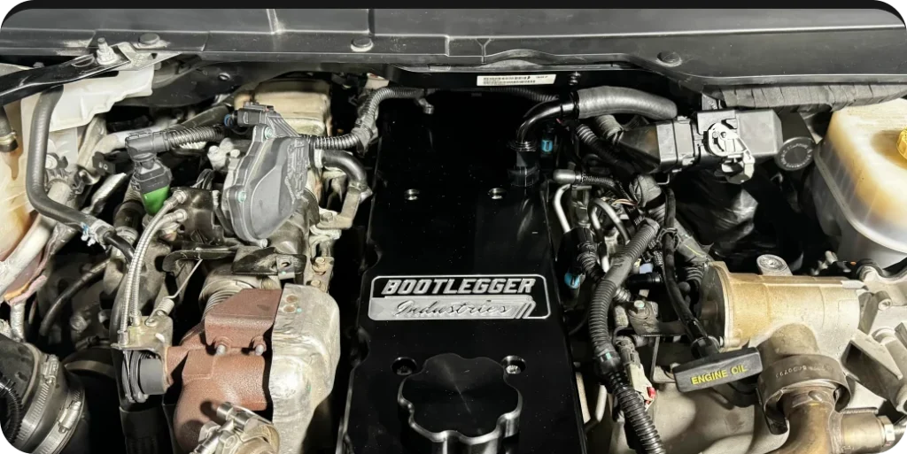 Close-up of a car engine with a "Bootlegger Industries" cover plate, and various hoses and components visible.